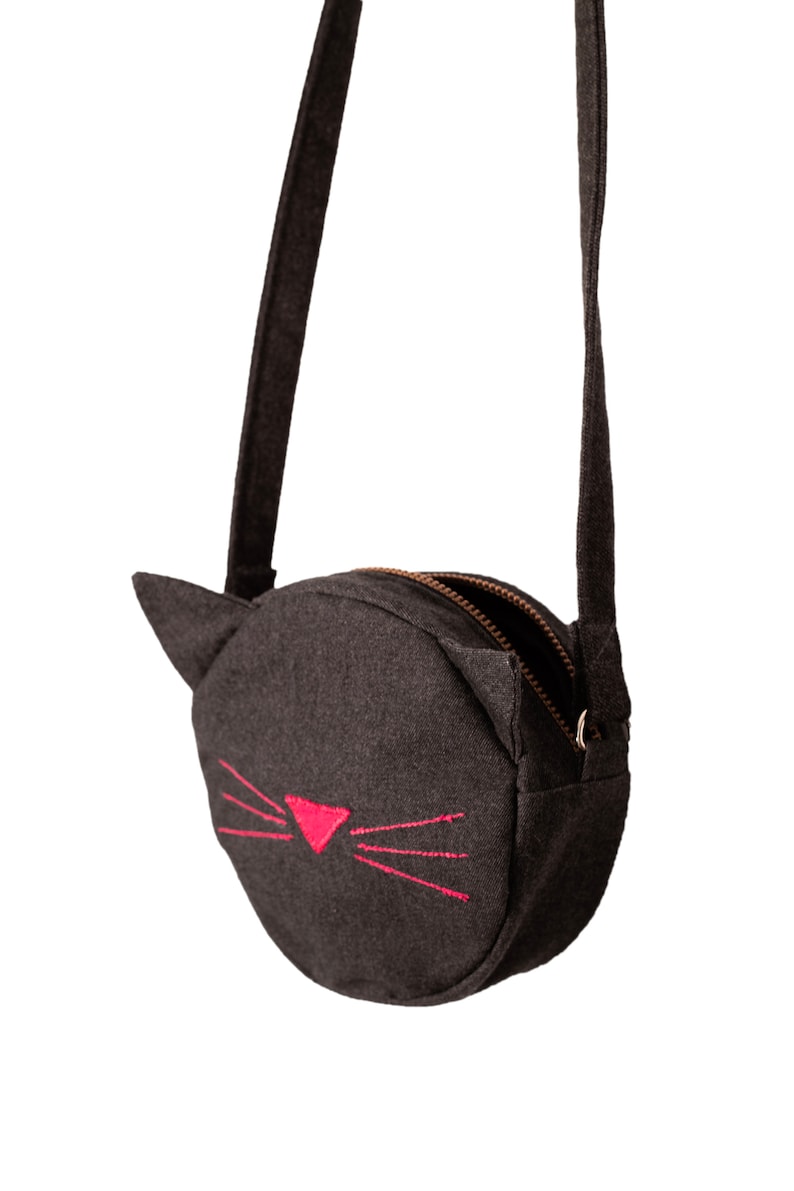 Puede incluir: Un peque&ntilde;o bolso de hombro redondo de fieltro gris con cierre de cremallera y una cara de gato bordada en rosa.