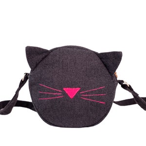 Puede incluir: Un bolso bandolera de fieltro negro con un dise&ntilde;o de cara de gato. El gato tiene bigotes bordados de color rosa y una nariz rosa.
