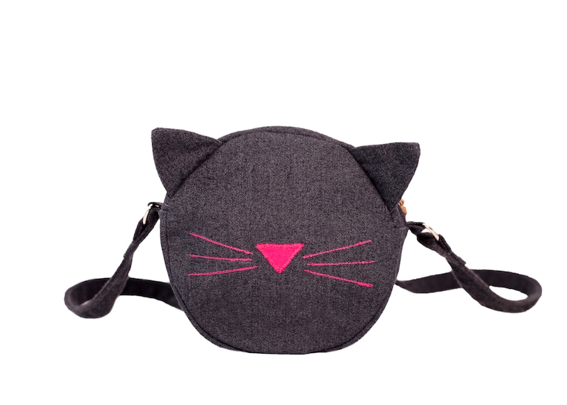 Puede incluir: Un bolso bandolera de fieltro negro con un dise&ntilde;o de cara de gato. El gato tiene bigotes bordados de color rosa y una nariz rosa.
