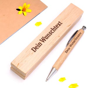 May include: A light-colored wooden pen case and a matching wooden ballpoint pen with silver metal accents. The case is engraved with the text "Dein Wunschtext" and the pen is engraved with the text "Dein Wunschname" in a dark font.