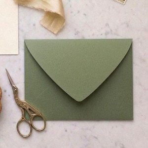 Può includere: Una busta verde oliva con patta a punta, su una superficie di marmo. Forbici dorate e un nastro color crema sono accanto alla busta. Un francobollo è visibile nell'angolo in alto a destra.