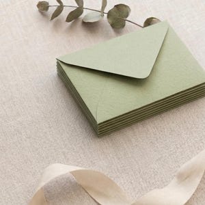 Pode incluir: Uma pilha de envelopes verde sálvia com um acabamento texturizado. Os envelopes estão dispostos sobre uma superfície de tom neutro, com um raminho de vegetação e uma fita creme adicionando elementos decorativos. Os envelopes são provavelmente para convites ou papelaria.