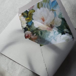 Könnte beinhalten: Ein hellgrauer Umschlag mit einer gerissenen, ausgefransten Kante, der teilweise geöffnet ist und eine Karte zeigt. Die Karte ist mit einem malerischen Blumenmuster bedruckt, das große weiße Pfingstrosen, grünes Laub und blaue Akzente zeigt.