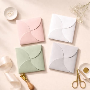 Op de afbeelding: Vier vierkante enveloppen in pastelkleuren: roze, wit, groen en grijs. De enveloppen hebben een unieke bloembladachtige sluiting. Accessoires zijn een klein schaaltje met ringen, een schaar, een lakzegel en lint, op een lichtroze ondergrond.