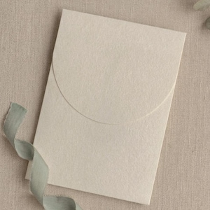 Puede incluir: Un sobre de invitación color crema con solapa curva, acompañado de una cinta verde claro. El sobre es de papel texturizado y la cinta está suelta. El fondo es una tela neutra y texturizada.