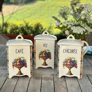 Puede incluir: Tres botes de cerámica vintage con ilustraciones de frutas y etiquetas que dicen "Café", "Riz" y "Chicoree". Los botes están sobre una mesa de madera con un fondo borroso de un campo verde y árboles.