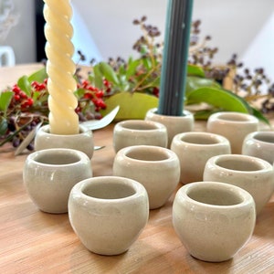 以下が含まれることがあります： 木製の表面に円形に配置された、10個の小さな白いセラミック製キャンドルホルダーのセット。ねじれた白いキャンドルがホルダーの1つに入っています。