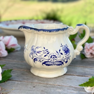 Könnte beinhalten: Eine weiße Keramik-Milchkanne mit blauem Blumenmuster. Die Kanne hat einen gewellten Rand und einen Henkel.