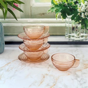 May include: A set of four pink glass teacups and saucers stacked on a white marble surface. The cups and saucers have a delicate, fluted design.