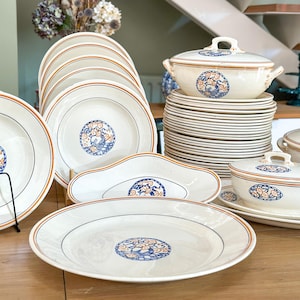 Peut inclure: Ensemble de vaisselle en céramique blanche avec des détails orange et bleus. L'ensemble comprend des assiettes, des bols et un plat de service couvert. Les assiettes ont un fin bord orange et un motif floral bleu et orange.