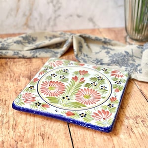 Puede incluir: Salvamanteles cuadrado de cerámica con borde azul y centro blanco con un diseño floral con flores rosas y verdes.