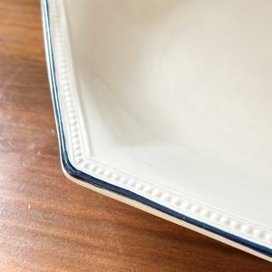 Pu&ograve; includere: Un piatto esagonale color crema caratterizzato da una texture a perline in rilievo lungo il bordo e da una sottile linea dipinta in blu scuro che ne definisce il contorno. Il piatto presenta una finitura liscia e lucida.