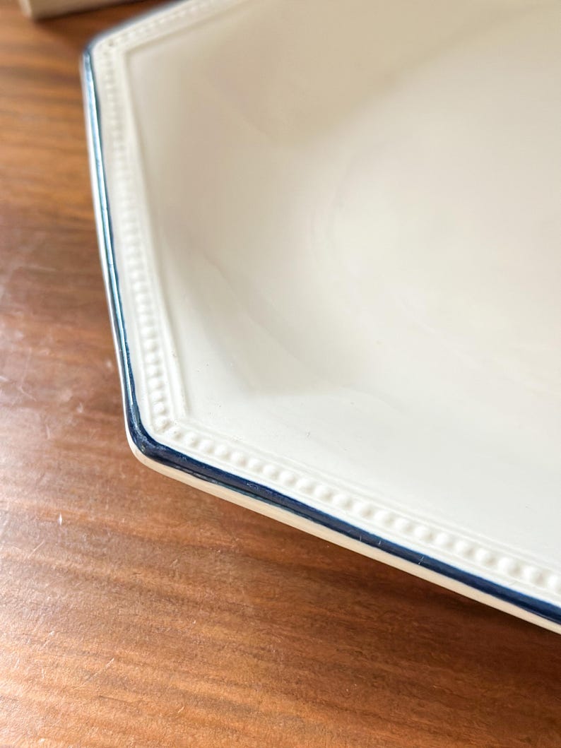 Pu&ograve; includere: Un piatto esagonale color crema caratterizzato da una texture a perline in rilievo lungo il bordo e da una sottile linea dipinta in blu scuro che ne definisce il contorno. Il piatto presenta una finitura liscia e lucida.