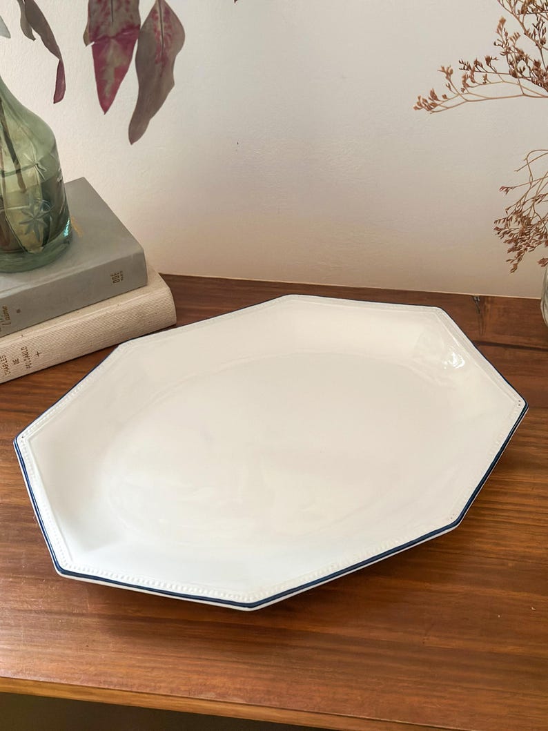 Pu&ograve; includere: Un piatto da portata ottagonale bianco con una finitura liscia e lucida e un sottile bordo blu scuro. Il piatto presenta un bordo leggermente rialzato con una delicata texture perlata, offrendo un design pulito ed essenziale per la presentazione.