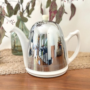 Peut inclure: Une théière vintage avec une partie supérieure brillante et argentée et une base en céramique blanche. Elle possède un bec verseur incurvé, une anse et un couvercle avec un bouton crème. Le design comprend une bande argentée autour de la base.