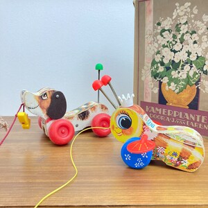 Può includere: Un giocattolo di legno vintage da tirare con un cane marrone e bianco e un'ape gialla e bianca con una corona. Il cane ha una ruota rossa e nera e l'ape ha una ruota blu e rossa. I giocattoli sono collegati da uno spago giallo.