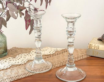 May include: Two clear glass candle holders with a tall, slender stem and a wide, circular base. Each holder features a series of decorative, rounded rings along the neck, providing a textured appearance to the smooth, transparent glass surface.