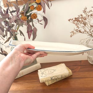 Pu&ograve; includere: Un vassoio da portata allungato in ceramica color bianco sporco, caratterizzato da una forma ottagonale distinta e da un sottile bordo blu scuro. Il vassoio presenta una base liscia e poco profonda con bordi angolati e un design neutro.