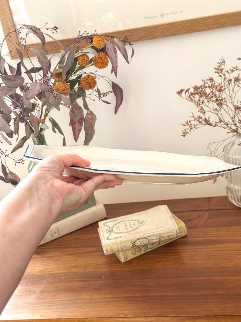 Pu&ograve; includere: Un vassoio da portata allungato in ceramica color bianco sporco, caratterizzato da una forma ottagonale distinta e da un sottile bordo blu scuro. Il vassoio presenta una base liscia e poco profonda con bordi angolati e un design neutro.
