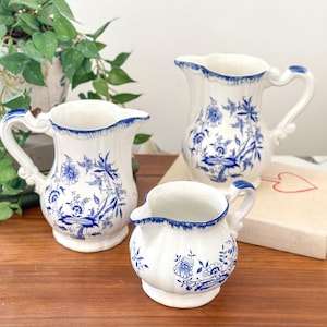 Può includere: Tre brocche in ceramica bianca con motivi floreali blu. Le brocche sono di diverse dimensioni e sono disposte su una superficie in legno.