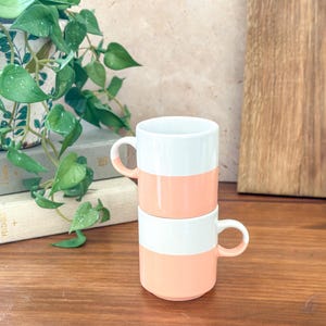 May include: Two stacked ceramic mugs. Each mug is white on top and coral-colored on the bottom, with a white handle. The mugs are on a wooden surface, with a plant and a wooden cutting board in the background.