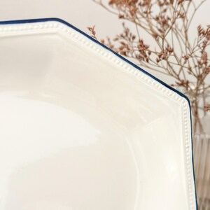 Pu&ograve; includere: Una ciotola in ceramica bianca di forma esagonale con un bordo blu scuro. Il bordo della ciotola presenta un dettaglio testurizzato a perline in rilievo che corre lungo tutto il perimetro, aggiungendo un sottile elemento decorativo alla superficie liscia e lucida.
