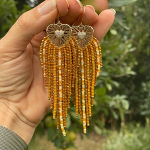 Könnte beinhalten: Goldfarbene Ohrringe mit herzförmigem Filigranmuster und einem kleinen Cluster aus Kristallen. Die Ohrringe haben lange Fransen aus kleinen, farbigen Perlen in Braun-, Beige- und Weißtönen mit goldenen Akzenten. Die Ohrringe sind zum Hängen gedacht.