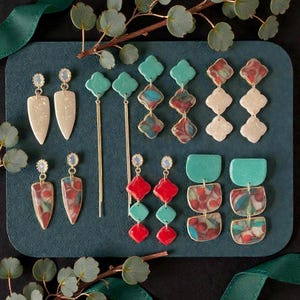 May include: A collection of handmade earrings in various designs and colors. The earrings include teardrop, geometric, and dangling styles, with colors like cream, teal, red, and marbled patterns. Each pair has a gold-tone metal accent and a decorative top.