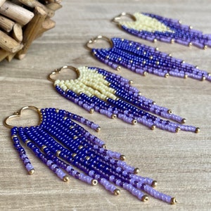 May include: Handmade beaded earrings with a heart-shaped gold hoop. The earrings feature a gradient of colors, starting with a cream-colored top, transitioning to deep blue, and ending with a light purple fringe. Gold beads accent the design.