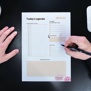 Può includere: Una persona sta scrivendo su un foglio di carta bianco con una penna nera. Il foglio ha un'agenda giornaliera stampata con sezioni per il blocco del tempo, i compiti più importanti del mattino e una sezione di scarico del cervello. Il testo "Today's agenda" è stampato nella parte superiore della pagina.