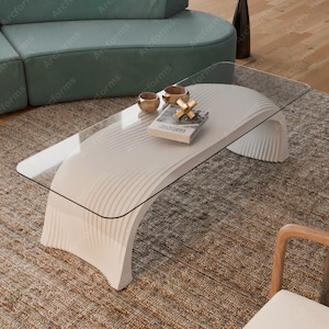 Puede incluir: Mesa de centro blanca con tapa de vidrio y un diseño curvo en forma de onda. La mesa está sobre una alfombra beige con un libro y dos tazas doradas en la parte superior.