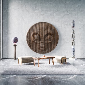 Op de afbeelding: Een houten sculptuur van een alien gezicht hangt aan een grijze muur, met een houten totempaal rechts en een ronde, grijs-rode bol op een voetstuk links. Twee houten banken en een houten salontafel staan voor een pluche wit tapijt.