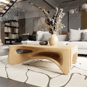 May include: A light brown, modern coffee table with a unique, layered design. The table is rectangular with curved cutouts and measures approximately 48 inches long. A vase with dried flowers and a decorative stone sit on top of the table, which is placed on a patterned rug.