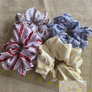 May include: Four fabric hair scrunchies featuring various patterns including ladybugs, floral motifs, and geometric designs. The scrunchies are arranged on a yellow plastic ruler with markings in inches, with the text Pinkish Sakura Studio visible.