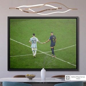 May include: A black framed canvas print of two soccer players, Lionel Messi and Kylian Mbapp&eacute;, shaking hands on a green soccer field. Messi is wearing a light blue and white jersey with the number 10 and Mbapp&eacute; is wearing a dark blue jersey with the number 10. The image is a close-up of the players from the waist up.