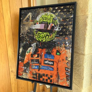 May include: Framed artwork featuring a portrait of a race car driver in a neon green and black helmet and orange racing suit. The artwork has a black frame and is displayed on a black easel. The background has a splatter paint effect.