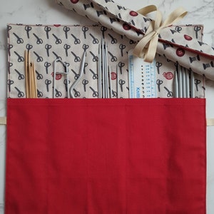 May include: A red fabric roll-up case with four compartments for storing knitting needles. The case has a beige fabric flap with a repeating pattern of scissors and buttons. The flap is rolled up and tied with a beige ribbon. A ruler with the word "KnitPro" is visible inside the case.