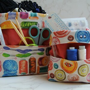 Op de afbeelding: Twee opbergmanden van stof met kleurrijke knoopmotieven. De grotere mand heeft een rode voering en bevat naai-accessoires, waaronder garen, naalden, scharen en pennen. De kleinere mand heeft een rode voering en bevat spoelen garen, een liniaal en een schaar.