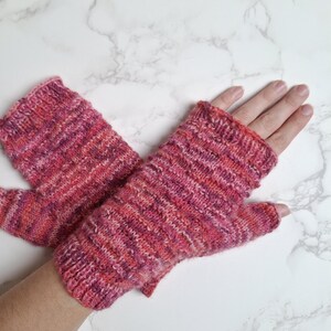 Könnte beinhalten: Ein Paar gestrickte Fingerhandschuhe in Rosa-, Rot- und Lilatönen. Die Handschuhe sind aus einem strukturierten Garn mit geripptem Bündchen und Daumenöffnung gefertigt. Die Handschuhe werden auf einer weißen Marmoroberfläche gezeigt.