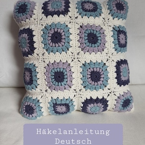 Könnte beinhalten: Ein gehäkeltes Kissen mit weißem Hintergrund und blauen, lila und grauen Granny Squares. Das Kissen hat den Text "Häkelanleitung Deutsch" unten.
