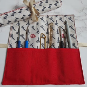 May include: A red fabric roll-up case with three compartments for storing knitting needles and crochet hooks. The case has a white fabric exterior with a repeating pattern of scissors and a ball of yarn.