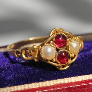 Könnte beinhalten: Ein goldener Ring mit roten Edelsteinen und Perlen, platziert auf einer samtigen Oberfläche in einer rot-blauen Schmuckschatulle. Der Ring weist aufwendige Filigranarbeiten auf.