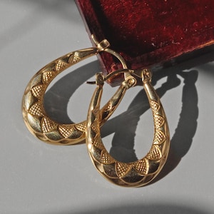 May include: A pair of gold-coloured hoop earrings with a textured, geometric design. The earrings are oval-shaped and have a clasp closure. They are displayed on a grey surface next to a burgundy velvet jewellery box.