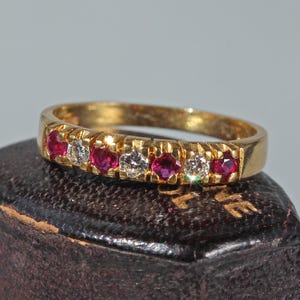 Puede incluir: Un anillo de oro con una hilera alterna de siete pequeñas piedras preciosas de corte redondo, que incluye cuatro rubíes rojos vibrantes y tres diamantes blancos transparentes, engastados en el canal metálico de la pieza.
