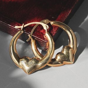 Puede incluir: Un par de pendientes de aro dorados con dijes en forma de coraz&oacute;n. Los pendientes se exhiben sobre una superficie gris claro con un fondo de madera rojo oscuro. Los pendientes est&aacute;n hechos de un material dorado brillante.