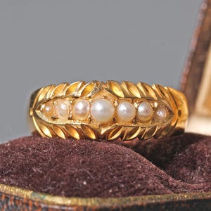 Op de afbeelding: Een gouden ring met een rij van vijf kleine witte parels gezet in een band versierd met een uitgesneden bladpatroon. De ring wordt gepresenteerd in een bruine, met fluweel gevoerde juwelendoos met een crèmekleurig binnendeksel.