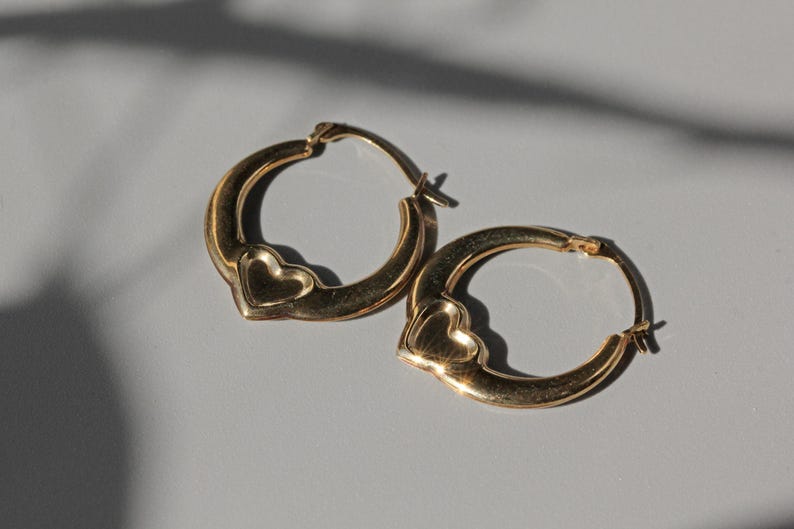 Puede incluir: Un par de pendientes de aro dorados. Cada pendiente presenta un dise&ntilde;o en forma de coraz&oacute;n en la parte inferior. Los pendientes se muestran sobre una superficie gris claro, con sombras proyectadas en el fondo.