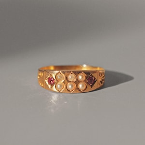 May include: A gold ring with a band decorated with a repeating pattern. The band is set with five white pearls and two red gemstones.