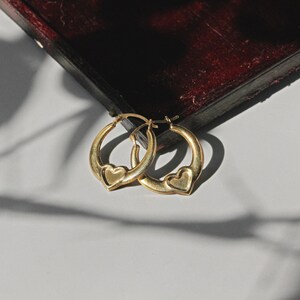 Puede incluir: Un par de pendientes de aro dorados, cada uno con un dije en forma de coraz&oacute;n. Los pendientes se exhiben sobre una superficie blanca, con una caja de madera oscura en el fondo. Son una joya.
