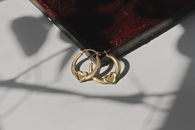 Puede incluir: Un par de pendientes de aro dorados, cada uno con un dije en forma de coraz&oacute;n. Los pendientes se exhiben sobre una superficie blanca, con una caja de madera oscura en el fondo. Son una joya.